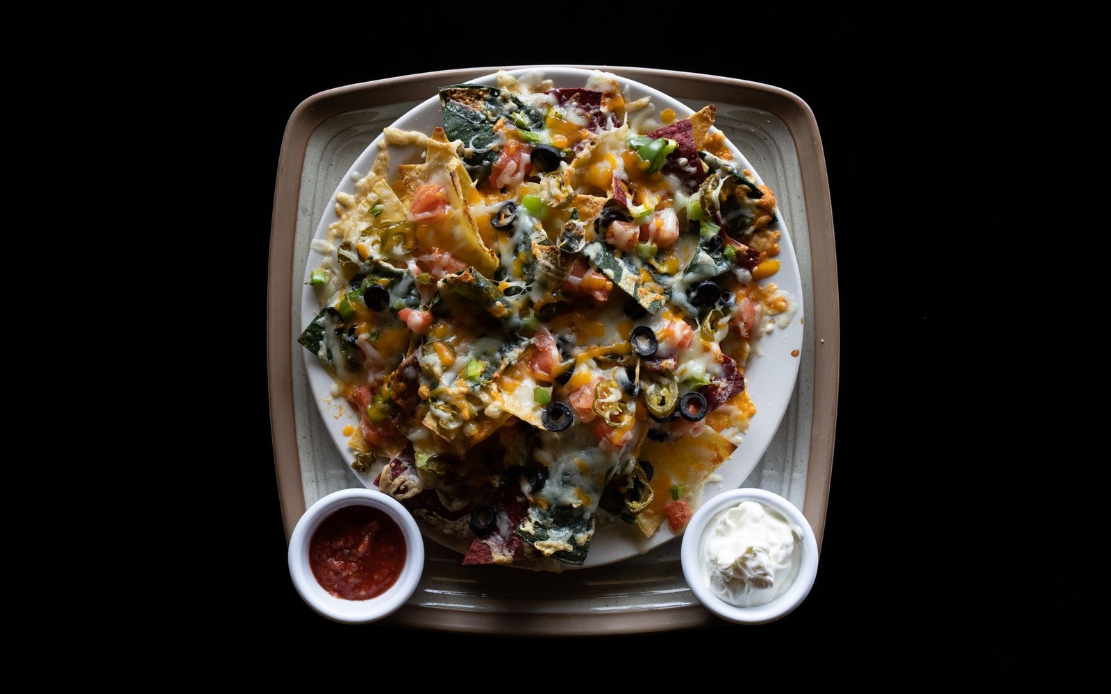 Plate of loaded nachos topped with melted cheese, olives, tomatoes and jalapeños, served with salsa and sour cream