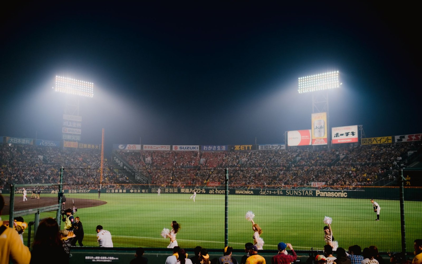 Floodlit baseball stadium at night with cheerleaders performing near the outfield fence
