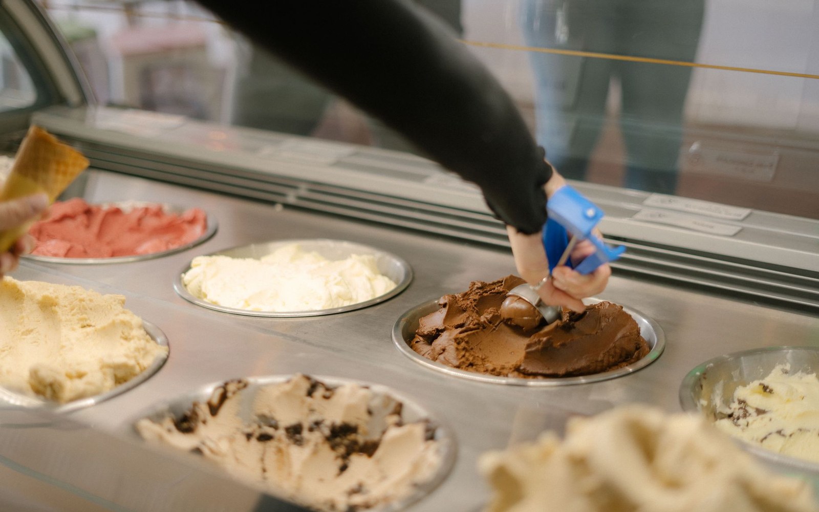 A server scoops chocolate gelato from a metal tray in a display case