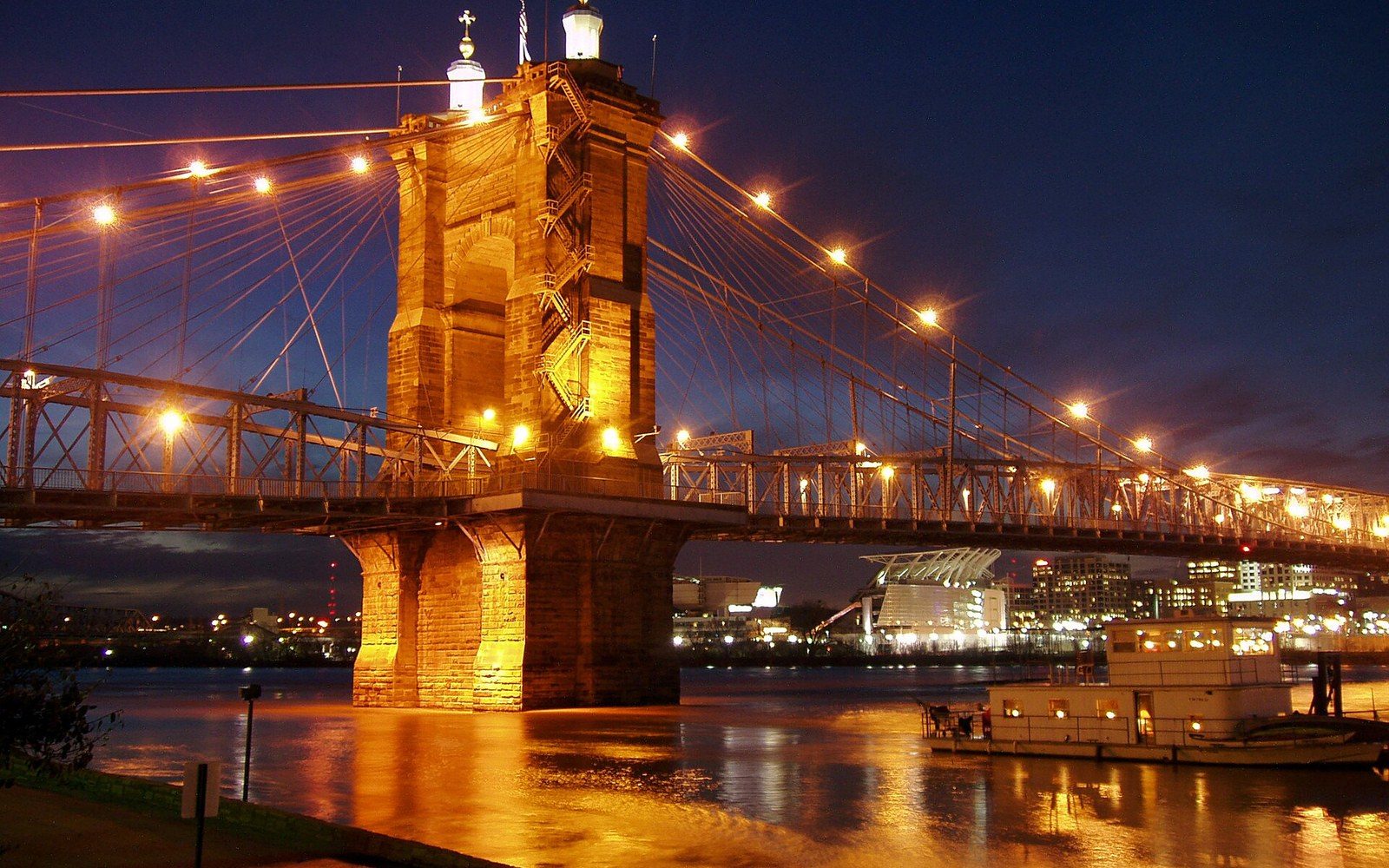 Suspension bridge tower illuminated at night over a river with a paddleboat below