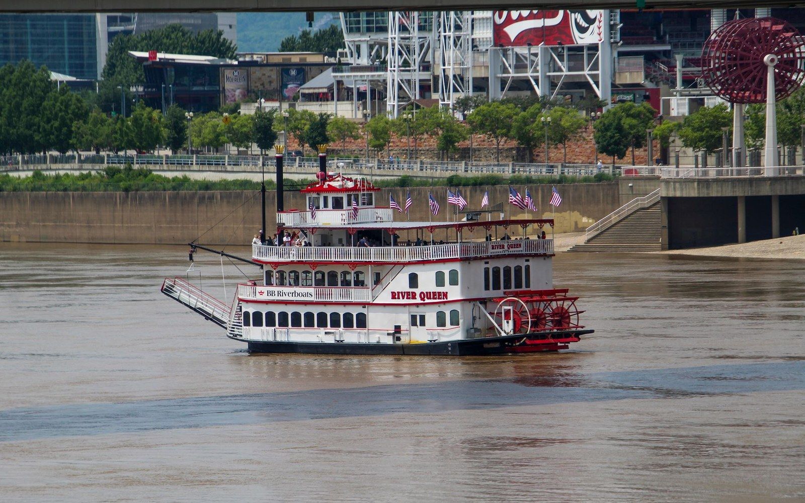 The River Queen paddlewheel boat cruises on a muddy river past a riverside cityscape